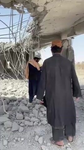 The video depicts a scene of extensive structural damage within a building, possibly the aftermath of a disaster or conflict. Several men are seen walking through the debris-filled interior.

The building's roof has largely collapsed, revealing a dense and chaotic web of twisted rebar and shattered concrete. Large chunks of concrete and other building materials litter the ground, forming a deep layer of rubble. The sky is visible through the gaping holes in the ceiling, suggesting a bright, clear day.

Three men are initially visible. One man in the foreground wears a dark tunic and a light-colored cap, walking away from the camera. Further back and to the left, another man in a light blue tunic and pants navigates the rubble. A third man, dressed in a light brown/beige tunic and a darker vest, is also seen moving through the wreckage. Later in the video, a fourth man, or possibly the foreground man from earlier, is shown from behind, wearing a white tunic and holding a camera up to his eye, appearing to be documenting the damage.

The overall scene is one of significant destruction, with exposed metal structures and broken concrete dominating the environment.