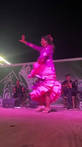 A flamenco dancer in a vibrant pink and purple polka dot dress with ruffled layers performs on a wooden stage under pink stage lighting. She is captured mid-twirl, her arms extended, and her dress flares out around her. In the background, two men are seated. One is playing a guitar, and the other is clapping his hands. A green banner with white graphics is visible behind the performers, and a dark, outdoor night sky is above. The overall atmosphere is of a live performance.