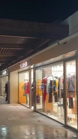 The video pans across a dimly lit outdoor promenade at night. There are several shopfronts with illuminated displays, showcasing clothing. One shop has a sign that appears to read "GUCCI". Further down the promenade, there is a cafe with patrons seated at tables. Plants in large pots are placed along the walkway. The ground appears wet, possibly from recent rain, and reflects the lights. The overall atmosphere is calm and relaxed.