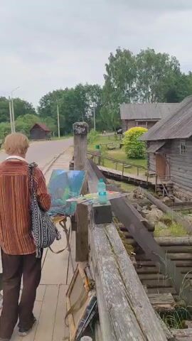 A person is standing on a wooden walkway, facing away from the camera. They are wearing a patterned bag over their shoulder and have their hands on a canvas that is set up on an easel. The canvas appears to be a painting of a landscape. To the right of the person, there is a water mill made of logs. The mill has a large wooden wheel that is partially submerged in the water. Pink flowers are growing in front of the mill. In the background, there are several wooden buildings and trees. The sky is overcast.