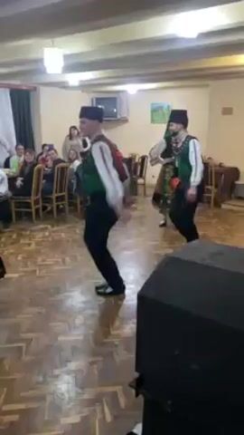 The video shows a group of people performing a traditional folk dance indoors. They are wearing colorful costumes that include vests, skirts, and headpieces. Some of the men are wearing hats. The dance is taking place on a wooden floor in a room that appears to be a banquet hall or event space. There are tables and chairs set up around the perimeter of the room, and several people are seated at the tables watching the performance. A large black speaker sits on the floor in the foreground. The room is lit by overhead lighting, and a television is mounted on the wall in the background.