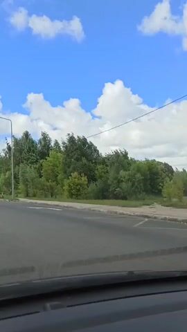 The video shows a view from inside a car as it drives down a road. The road has multiple lanes and is lined with trees and streetlights. There are other cars on the road, and in the distance, there are some tall buildings. The sky is blue with white clouds. There is a yellow electric scooter parked on the side of the road.