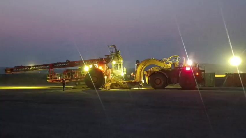 A large, yellow mining vehicle with an extended arm is stationary on a dirt or gravel surface. The sky is dark, suggesting it is either dawn or dusk. The vehicle is illuminated by its own lights, which are bright and cast a yellow and orange glow on the ground and surrounding area. There are also bright white lights illuminating the scene from the right. Two individuals are visible near the vehicle. One is standing closer to the front of the vehicle, and another is further away, near the middle of the vehicle. The vehicle appears to be a piece of heavy-duty mining equipment, possibly a drill or loader. The overall impression is of an industrial setting at a time of low natural light.