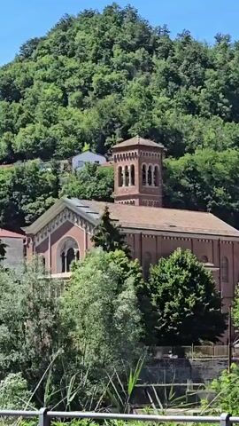 The video shows a brick church with a bell tower, nestled in a lush green hillside. Trees and foliage surround the building, and a clear blue sky is visible. The scene is peaceful and idyllic.