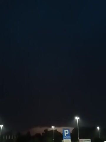 The video captures a dark, cloudy sky, possibly indicating an approaching storm. Several streetlights are visible, illuminating the lower part of the frame. In the distance, there are trees and what appear to be buildings or structures. A parking sign is also present. The overall atmosphere is dim and foreboding.