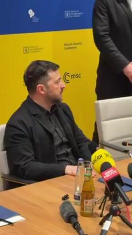 In this video, a man, likely Volodymyr Zelenskyy, is seated at a table in front of a yellow backdrop featuring logos and text indicating a Munich Security Conference and the Office of the President of Ukraine. He is dressed in a black shirt and jacket. Several microphones from news organizations like "YHYAH" and BBC News are positioned on the table in front of him. A bottle of water and a bottle of an amber-colored beverage are also on the table. A portion of another person, dressed in black, stands behind and to the right of Zelenskyy. Zelenskyy is speaking and gesturing with his hands, suggesting he is giving an interview or making a statement.