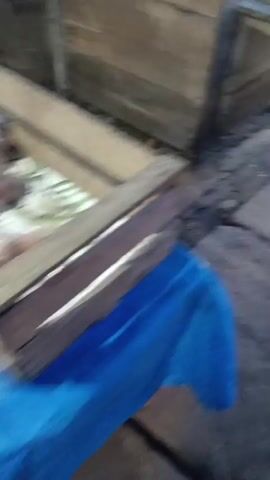 The video shows a ground-level view of a pathway made of square concrete tiles. To the left, there is a raised wooden structure with some debris underneath. The pathway continues to the right, where it appears to lead to an area with stalls or market stands. In the distance, a person in light-colored pants is partially visible. There are also various objects, including what appear to be containers and goods on display, though they are blurry and difficult to identify clearly. The lighting suggests an outdoor environment, possibly during the day.