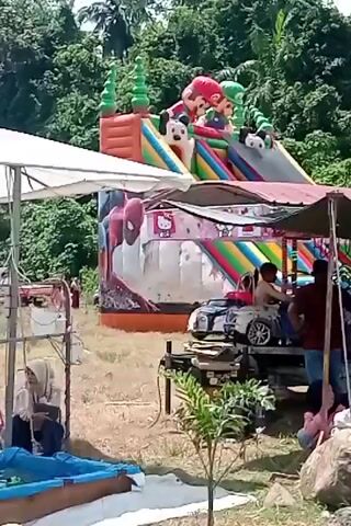 The video shows an outdoor amusement area with an inflatable slide in the background and a row of small amusement rides in the foreground. The inflatable slide is colorful and features characters resembling Mickey Mouse and Super Mario. Several children are visible on the slide, with some sliding down and others at the top.

In the foreground, under a canopy, are several small, electric-powered car rides arranged in a line. Children are seated in these cars, and some appear to be in motion. Several adults are also present, some standing and observing, while others are seated on chairs near the rides. One woman in a white headscarf is looking at her phone. The ground is covered in grass and some large rocks. The area is surrounded by lush green trees, suggesting a natural or park-like setting. The overall atmosphere appears to be a daytime family outing or a local fair.