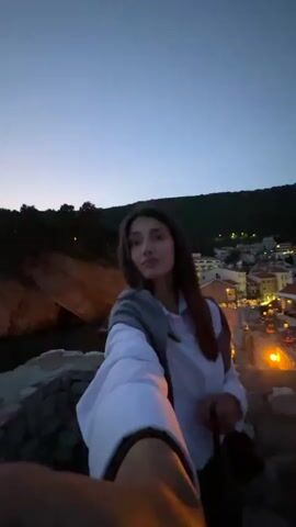 The video shows a young woman taking a selfie with a background of a coastal town at dusk. She is standing on a stone wall, holding the camera out towards herself. She has long brown hair, fair skin, and is wearing a white shirt with a grey jacket. She smiles slightly at the camera. Behind her, there is a rocky cliff on the left and a view of the town on the right. The town has white buildings with red roofs and is illuminated by streetlights. The sky is a deep blue, indicating it is dusk. The video is shot from a low angle, giving the woman a slightly elevated position.