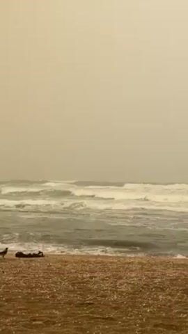 The video shows a hazy view of an ocean scene from the perspective of a beach. The sky is overcast, with a yellowish-grey tint obscuring details. The ocean extends horizontally across the frame, with choppy waves moving towards the shore. The beach in the foreground appears composed of small pebbles or sand. The overall scene conveys a sense of atmospheric obscurity and a potentially rough sea condition.