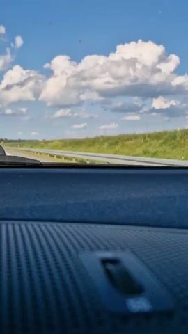 The video shows a view from inside a car looking out the windshield. The dashboard is in the foreground, with a textured surface. The road ahead is visible, with a guardrail on the left and a grassy embankment on the right. The sky is blue with white, puffy clouds. There are a few small red dots visible on the windshield, and a yellow sign with a black symbol is visible in the distance.