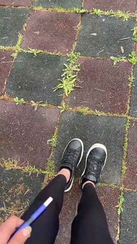 The video begins with a first-person perspective of a person walking on a playground with rubberized ground tiles. The person is wearing black leggings, black sneakers, and is holding a blue pen. The camera pans up to show a grassy area with a few children's toys, including a green toy tractor and a yellow toy scooter. Further in the background, there are several park benches under a large tree. The camera continues to pan, revealing a wider view of the playground, which includes a swing set, a merry-go-round, and a small wooden gazebo. The sky is cloudy and appears to be overcast. The overall environment suggests an outdoor recreational area, possibly a park or a school playground.