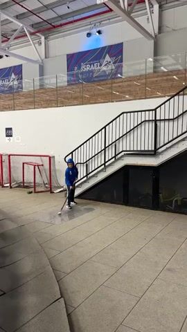 The video shows a young person in a blue jacket and helmet playing hockey indoors. They are on a tiled floor with a hockey stick and a puck. There is a hockey net in the background, as well as stairs and banners. The scene appears to be an indoor sports facility. The camera pans to the right, revealing more of the facility, including a ladder, another hockey net, and some equipment. The lighting is bright and consistent throughout the video.