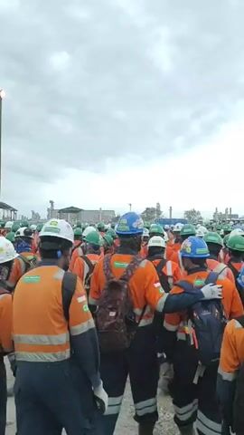 The video shows a large group of people wearing orange work shirts and hard hats, some with backpacks, gathered outdoors. They appear to be workers on a construction site or industrial facility, with a tower emitting a flame in the background. The sky is overcast. The individuals are facing away from the camera, suggesting they are either waiting for instructions or participating in a gathering. Some people are wearing blue hard hats, while others have green or white hard hats. The overall impression is a workforce assembled, possibly for a morning briefing or a shift change.