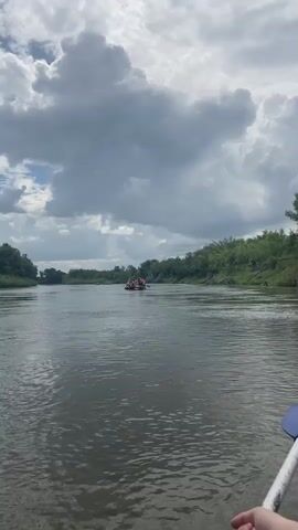 A group of people in orange life vests are on a raft in a wide river. The raft is moving downstream. The riverbanks are lined with trees. The sky is cloudy.