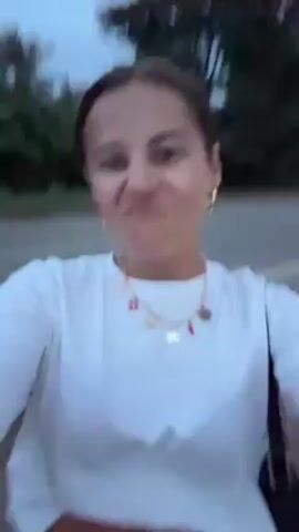 A person with medium-brown hair pulled back into a bun, wearing a white cropped t-shirt, blue jeans, and a colorful necklace is holding a camera and filming themselves from a low angle. They are standing on a street or paved path. They make a face, sticking out their tongue. The background is blurry but appears to be trees and possibly other buildings. The lighting suggests it might be dusk or early evening.