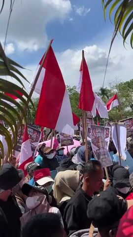 A crowd of people are gathered outdoors, holding Indonesian flags and signs. The flags are red and white, and many of the signs have text in Indonesian. Some people are wearing masks. In the background, there is a stage with two men, one of whom is speaking into a microphone. The sky is blue with some clouds. Palm fronds are visible in the foreground, partially obscuring the view. The overall impression is of a political rally or demonstration.