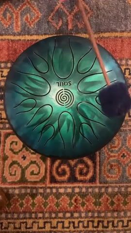 A top-down shot shows a steel tongue drum with a green and blue iridescent finish placed on a patterned rug. The drum has a circular maze-like design in the center with the word "SOUL" written above it. The drum is being played by someone's hands, which enter the frame from the top right, holding two mallets. The mallets strike different sections of the drum, producing musical notes.