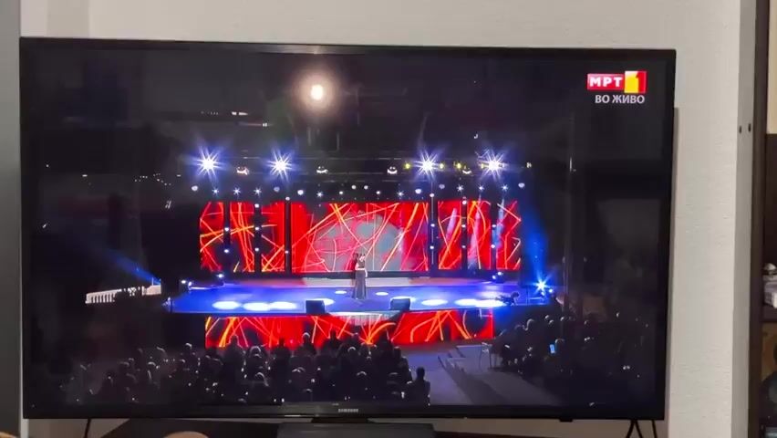 The video displays a television screen. The first few seconds show a live broadcast of a performance on a stage with bright red and blue lights and a crowd of people watching. A singer stands in the center of the stage. The MPT 1 logo is visible in the top right corner of the screen. The scene then transitions to a colorful graphic interface with multiple smaller images displayed in squares and rectangles against an orange background. In the center, there is a dark gray square with a stylized ladybug logo inside. One of the smaller images shows the MPT 1 logo. The transition is quick, and the graphic interface remains on screen for the rest of the video.
