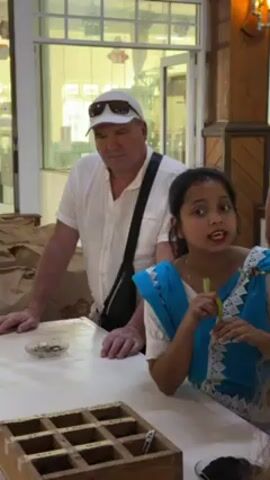 A young woman in a blue sari with white lace stands behind a white counter, gesturing with her right hand and speaking with her mouth open. Behind her and to her left stands an older man in a white short-sleeved shirt, a white baseball cap, and sunglasses. He has a black bag slung across his chest. He looks at the woman while she speaks and then scratches his chin with his right hand. In front of the pair is a wooden tray with nine compartments, each containing what appears to be tea leaves. Small round dishes containing tea leaves also sit on the counter. Large brown paper bags are visible on the counter behind the man. A glass window and door are behind the man. A wooden wall with a decorative light fixture is to the right.