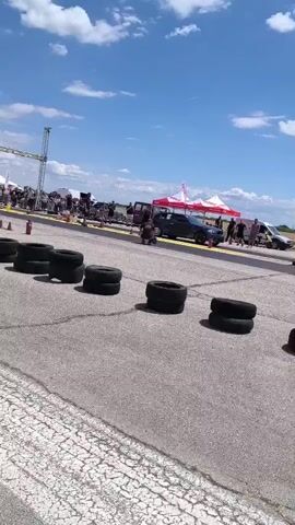 The video shows a drag race event on a sunny day. Several cars are visible on the asphalt track. In the foreground, there are stacks of tires. In the background, there are tents and people watching the race. The sky is blue with white clouds. The camera pans across the scene, capturing the cars as they race down the track.