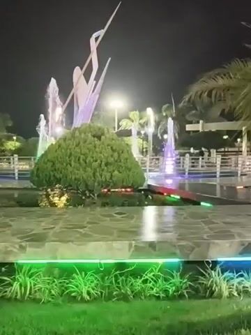 The video depicts a night scene in a park-like area.  A fountain feature is centrally located, surrounded by stone pavement and low landscaping. The fountain is illuminated with colored lights and has several jets of water spraying upwards. A modern sculpture is positioned near the fountain. Palm trees are visible in the background and periphery. Lampposts provide additional lighting. The general atmosphere is well-lit and appears tranquil.