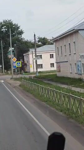 The video shows a street scene from a moving vehicle. The perspective is from the driver's seat or passenger seat of a car. A pedestrian is visible crossing the street on a crosswalk. There are several buildings, trees, and streetlights along the road. Traffic signs, including a pedestrian crossing sign and a medical emergency sign, are visible. Several vehicles, including cars and trucks, are driving on the road. The weather appears to be overcast.