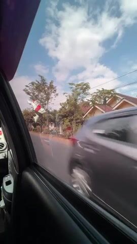 The video shows a view from inside a vehicle, looking out the passenger-side window. The window frame and part of the car interior are visible on the left. The road runs diagonally from the bottom left to the upper right. On the right side of the road, there are houses, some with pink walls and red roofs, surrounded by trees. There are also power lines visible. The sky is blue with scattered white clouds. A white object with red and white stripes is visible in the distance, hanging from what appears to be a pole or wire. The lighting suggests it is daytime.