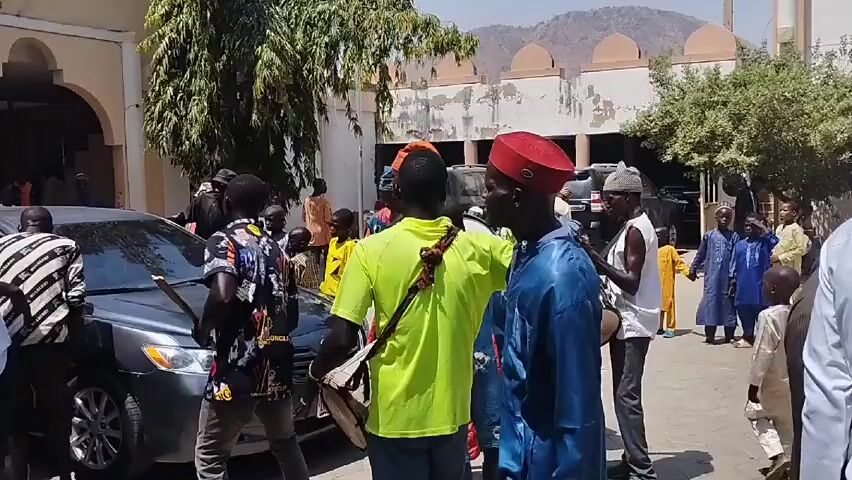 The video depicts a lively outdoor scene with a crowd of people gathered, some dressed in traditional African attire. The environment features a building with an archway, several parked cars, trees, and in the distance, a mountain range is visible under a bright sky. The people are interacting, some playing musical instruments like drums and wind instruments, while others are watching or walking around. There is a sense of celebration or an event taking place, as the music and gathering suggest a social or cultural occasion.