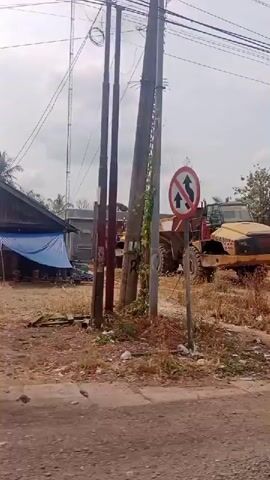 The video begins with a view of a yellow construction vehicle, possibly a bulldozer, parked on a dirt lot with debris and some building materials around it. In the background, there are trees and houses. The camera then pans to the right, showing a green dump truck driving on a dirt road. Next, the camera captures a side view of a modern house with a gate and a motorcycle parked in front. The video continues to show a motorcycle with two people riding on it passing by the same house. Finally, the camera shows a traffic light pole with a functional traffic light and a gate to a property. The sky is overcast throughout the video.