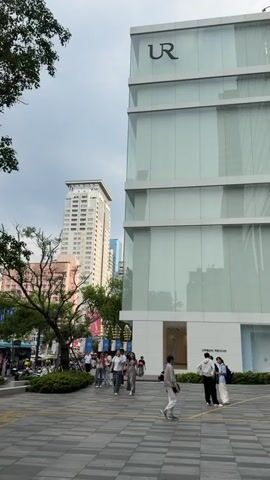 The video pans across a modern urban street scene. In the foreground, a paved plaza with a checkered pattern is visible, with people walking and standing. To the right, a large, multi-story building with a glass facade dominates the view. The building has a prominent "UR" logo at the top and "URBAN REVIVO" signage near the bottom. Several people are walking in front of this building, some singly and some in small groups. To the left of the glass building, there is a large, dark advertising screen displaying text and images, with more shops visible below it. Further to the left and in the background, a tall, light-colored skyscraper with many windows stands out against a cloudy sky. A leafy tree with pinkish flowers is situated between the paved plaza and the advertising screen. The overall atmosphere appears to be that of a bustling commercial district.