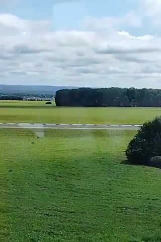 The video is an external shot looking out at a rural landscape. In the foreground, a tall, gray pole with a black number "9" is visible on the left side of the frame. Beyond the pole, there is a vast green field with scattered yellow flowers. A highway with a white and black barrier runs horizontally across the middle ground. In the background, there are more green fields, a cluster of trees, and a distant village with small houses. The sky is blue with scattered white clouds. The overall impression is a scenic, undeveloped area.