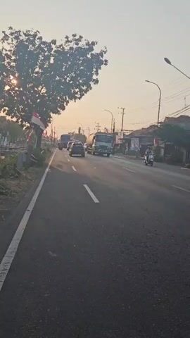 The video shows a road scene at sunrise or sunset, with the sun low in the sky casting a warm glow. The camera is mounted on a moving vehicle, likely a car or motorcycle, traveling along the right side of a two-lane road. Several vehicles are visible, including cars, trucks, and motorcycles, all traveling in the same direction. Some buildings and trees line the side of the road, and there are utility poles and wires overhead. Indonesian flags are visible on the side of the road, suggesting the setting is in Indonesia. The overall atmosphere is calm and mundane, depicting everyday traffic on a semi-urban road.