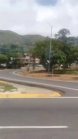 The video captures a view from a moving vehicle, looking out at a road and a landscape. The road appears to be paved with asphalt and has yellow and white markings. To the right of the road, there is a strip of dirt and grass with a yellow curb. Further to the right, there are trees and some buildings in the background. The buildings are a mix of residential and commercial structures, some of which appear to be multi-story. There are also signs visible on some of the buildings. The landscape is hilly, with vegetation covering the slopes. The sky is visible above, with some clouds present. There are also utility poles and wires visible in some parts of the frame. The overall scene suggests a semi-urban or suburban environment.