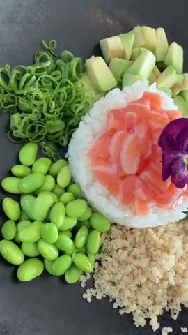 A close-up overhead shot shows a poke bowl being assembled. The bowl is divided into sections containing white rice topped with raw salmon and a purple flower, cooked quinoa, bright green edamame, diced avocado, and green seaweed salad. A white ramekin of dark brown sauce is tilted to pour the sauce over the salmon and rice. The background is dark, likely a plate or table. The lighting is bright, highlighting the vibrant colors of the food.