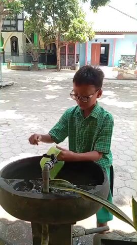 A young boy wearing glasses and a green checkered shirt is pouring water from a light green plastic container into a circular basin, which appears to be a water fountain. The fountain is outdoors, on a paved area with a hexagonal tile pattern. Behind the boy, there is a building with light-colored walls and a green roof, and a large tree. To the left of the building, there is a basketball hoop. The boy is facing the fountain and is in the process of emptying the container. The water is flowing from the container into the fountain, and there is a visible stream of water coming from a faucet in the fountain. There are some plants visible in and around the fountain. The overall environment suggests a schoolyard or a similar public outdoor space.