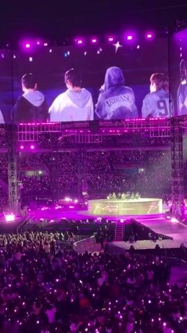 The video shows a K-pop performance in a stadium. A large video screen shows five men singing into microphones. Below the screen is a stage where the same five men are standing. The stadium is filled with people who are holding up small lights. The overall lighting is purple.