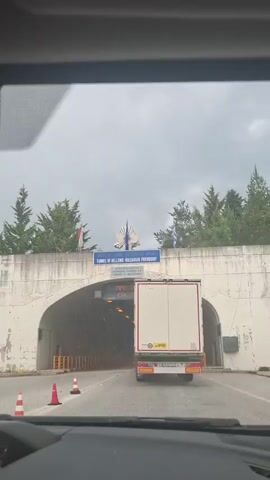 A white truck is driving through a tunnel. The tunnel is illuminated by yellow lights. The truck is in the center of the tunnel, and there are yellow barriers on either side of the road. The sky outside the tunnel appears to be cloudy. There is a sign above the entrance to the tunnel that reads "TUNNEL OF HELLENIC-BULGARIAN FRIENDSHIP".