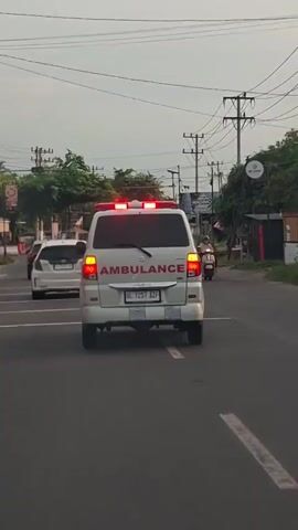 The video shows the rear of a white ambulance driving on a road in Cotbadatunong, Indonesia. The ambulance has red flashing lights on its roof and the word "AMBULANCE" printed in red letters on its back. The license plate reads "BL 1257 AZF". A white car is visible in front of the ambulance, and a person on a scooter is driving past on the right side of the road. In the background, there are trees, utility poles, and buildings, suggesting an urban or suburban setting. The sky is overcast, indicating it might be late afternoon or early evening. The overall scene depicts normal traffic conditions with an emergency vehicle in transit. There is also a traffic light visible on the left side of the image. A sign with the word "WADIMOR" is visible in the background.