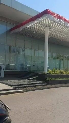 A motorcycle rider, seen from behind and from the right, is traveling on a paved road past a modern building with a glass facade and a covered entrance decorated with red and white fabric. The building has large glass panels, some of which appear frosted or tinted. There are stairs leading up to the entrance, and planters with greenery are situated along the front of the building. The sky is clear and blue, indicating a sunny day. The motorcycle is dark in color, and the rider is wearing a black helmet and a dark jacket. Another motorcycle is partially visible in the first second of the video, to the left of the frame.