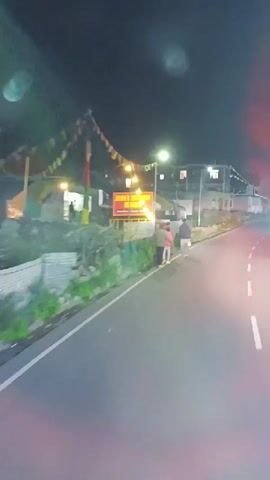 A nighttime video shows a paved road next to a grassy embankment. Several people walk along the side of the road. On the embankment, there are several signs. One sign, illuminated by lights, reads "ZION HIGHWAY DINER BREAKFAST | LUNCH". Prayer flags are strung across wires overhead. Buildings with illuminated windows are visible in the background. The scene appears to be in a town or village.