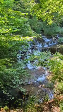 A narrow stream cascades over a series of small rocky ledges, forming miniature waterfalls. Lush green foliage, with sunlight filtering through, frames the scene. The water is clear and appears shallow, with white foam and turbulence visible where it tumbles over the rocks. The background consists of more trees and vegetation, suggesting a natural, wooded environment. There are no people or man-made objects visible. The perspective is from slightly above and to the side of the stream, looking down its length.