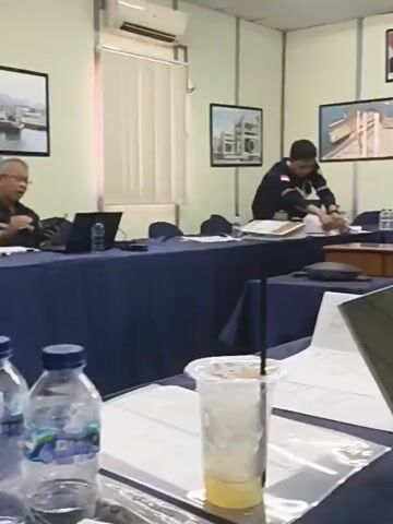 A video clip shows a meeting room. A man is seated at a table, looking at papers. Another person is visible in the background. The room has a whiteboard, pictures on the wall, and several chairs and tables. The tables are covered with blue tablecloths. There are laptops and papers on the tables. A sign on the wall reads "Bakrie Construction". The lighting is bright, and the overall atmosphere appears to be formal.