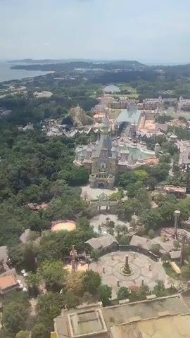 The video shows an aerial view of a theme park, with a large castle-like structure as the central focal point. The park features various buildings and attractions, including what appears to be a water park area with slides and pools. Lush green trees surround the park, and in the background, there is a view of a coastline with some buildings in the distance.  The sky is bright with some clouds. The video seems to be taken from a high vantage point, potentially a tall building within the park or a nearby structure.