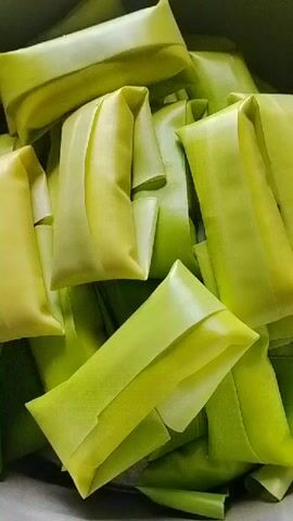 A close-up shot shows a pile of green food items, possibly wrapped in banana leaves. The items are rectangular and have a folded appearance. The lighting suggests an indoor setting, and the focus is entirely on the food. There are no people or other objects visible in the frame.