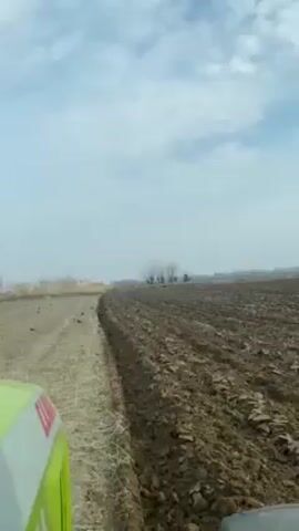 The video shows a view from the inside of a vehicle, presumably a tractor, moving across a field.  On the left is an area of undisturbed dry vegetation and to the right, freshly plowed brown earth.  In the distance are some bare trees on the horizon. The sky is blue with scattered white clouds.  The hood of the tractor is visible on the left edge of the frame, showing part of the word "CLAAS." The day appears bright.