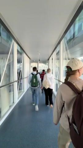 In a well-lit airport jet bridge or connecting walkway, several individuals are seen walking away from the camera. The person in the foreground wears a beige long-sleeved shirt, light-colored pants, a cream baseball cap, and a brown backpack. Ahead of them, a man with a green backpack and blue jeans walks alongside a woman carrying a white handbag. Other people are further ahead in the walkway. The walkway features large glass windows on the right side, a white wall on the left with a metal handrail, and a blue floor. Ceiling lights and security cameras are visible overhead. Around the 0:03 mark, a sign on the left wall with Chinese characters and the English word "Arrivals" is visible, along with an arrow pointing forward. A green arrow also marks the path on the floor. The scene suggests passengers are moving through an airport, likely after disembarking an aircraft or transferring between terminals.