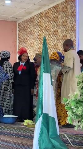 A group of people are gathered in what appears to be a church or community hall. A Nigerian flag stands prominently in the foreground. A woman in a black suit, red hat, and red scarf is speaking. Several other people in traditional attire are around her. There is a lectern with a glass top on the right side of the frame. The room has patterned wallpaper and a carpeted floor.
