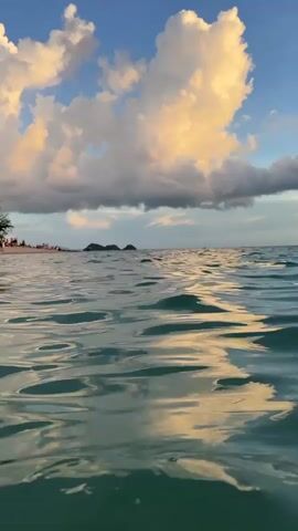 This video shows a low-angle view of the ocean. The water is calm with gentle ripples reflecting the sky. In the distance, there are two small islands or large rocks with some vegetation. The sky is filled with dramatic clouds that are illuminated by the setting sun, casting warm golden and orange hues. A few distant figures can be seen on the water, possibly swimming or on small boats. The overall atmosphere is serene and picturesque, characteristic of a sunset over the ocean.