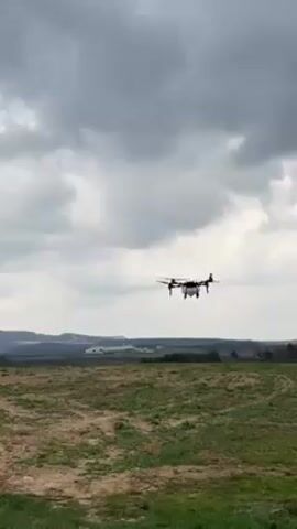 The video shows a quadcopter drone hovering over a field. The drone is black with white tanks underneath. The field is green and brown with some bare patches. In the background, there are hills and a cloudy sky. Two people are speaking in the background. The drone appears to be preparing to land.